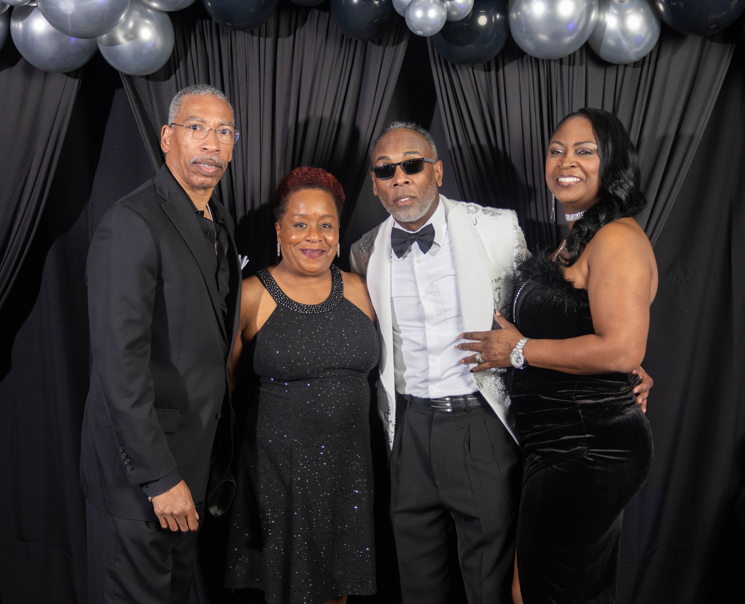 Four elegantly dressed people posing together at a formal event with black and silver balloon backdrop.
