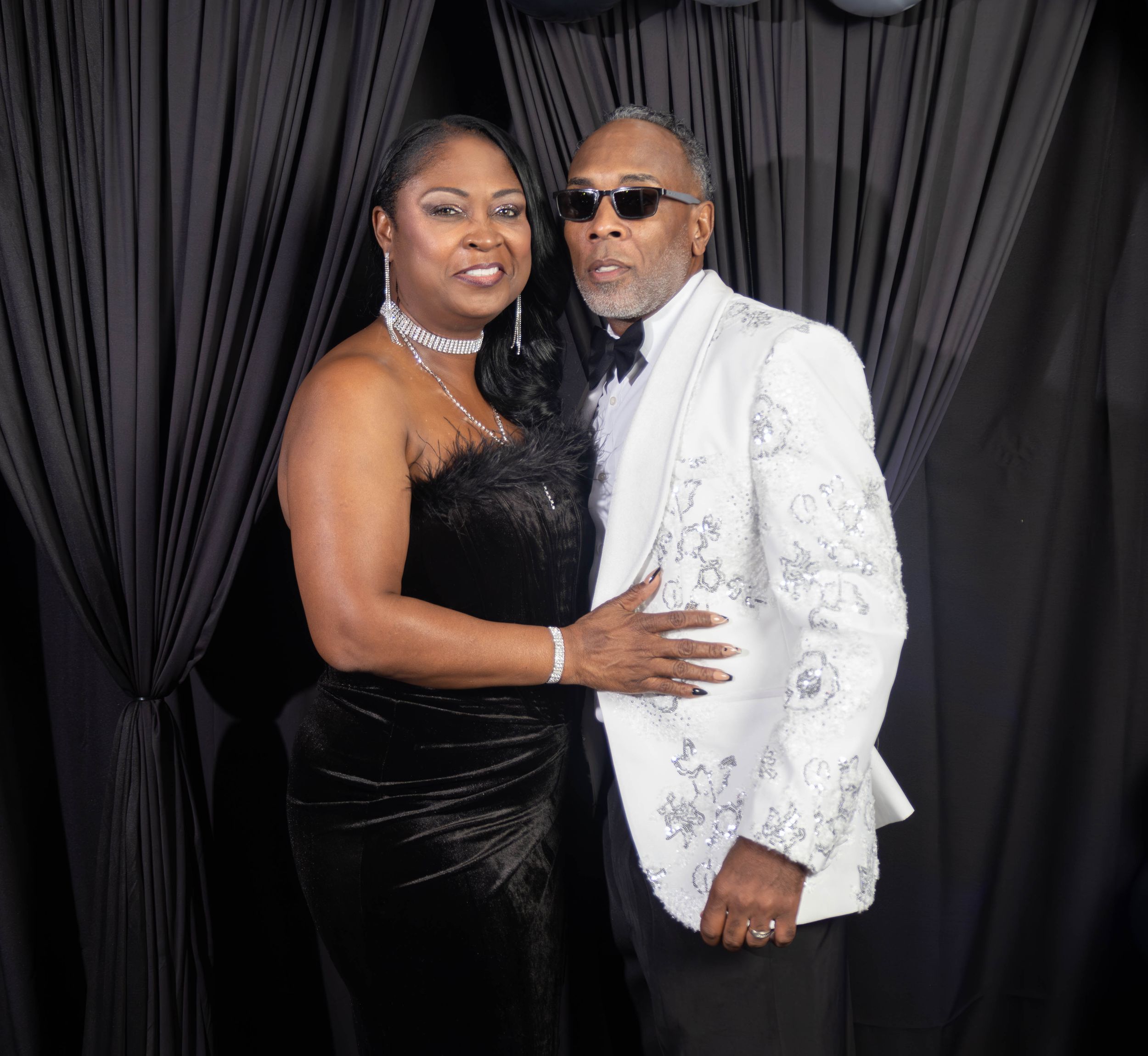 Elegant couple posing at formal event, woman in black gown and man in white embroidered tuxedo jacket.