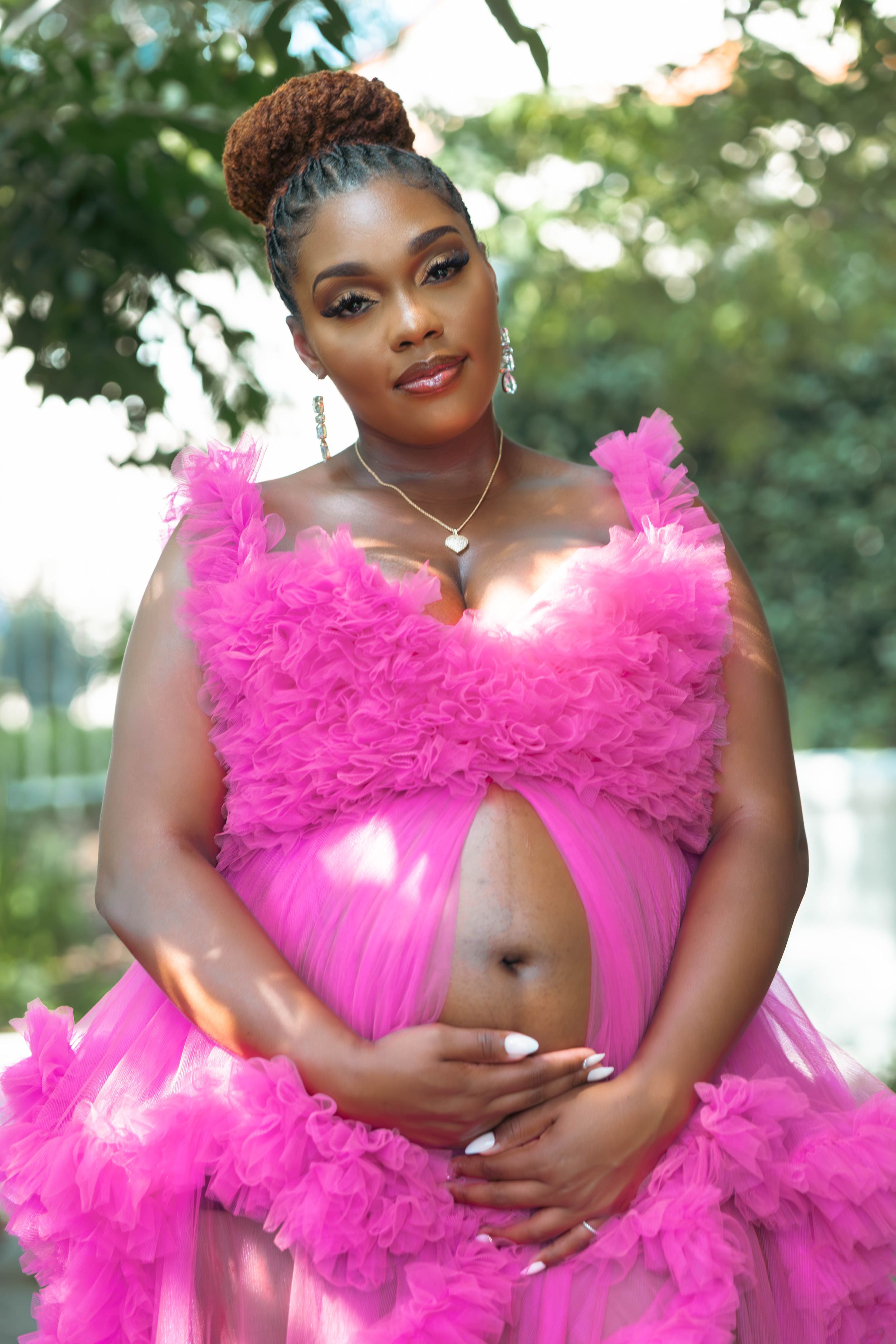 Pregnant woman in pink ruffled tulle maternity gown cradling baby bump outdoors among lush green trees.