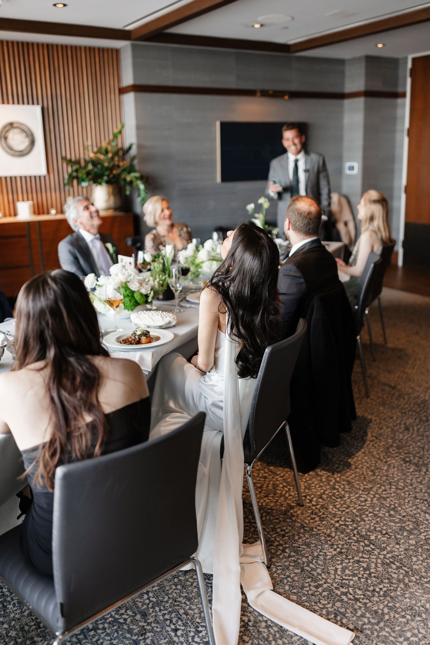 Elegant wedding reception with guests seated at a long table, listening to a toast in a modern dining room.