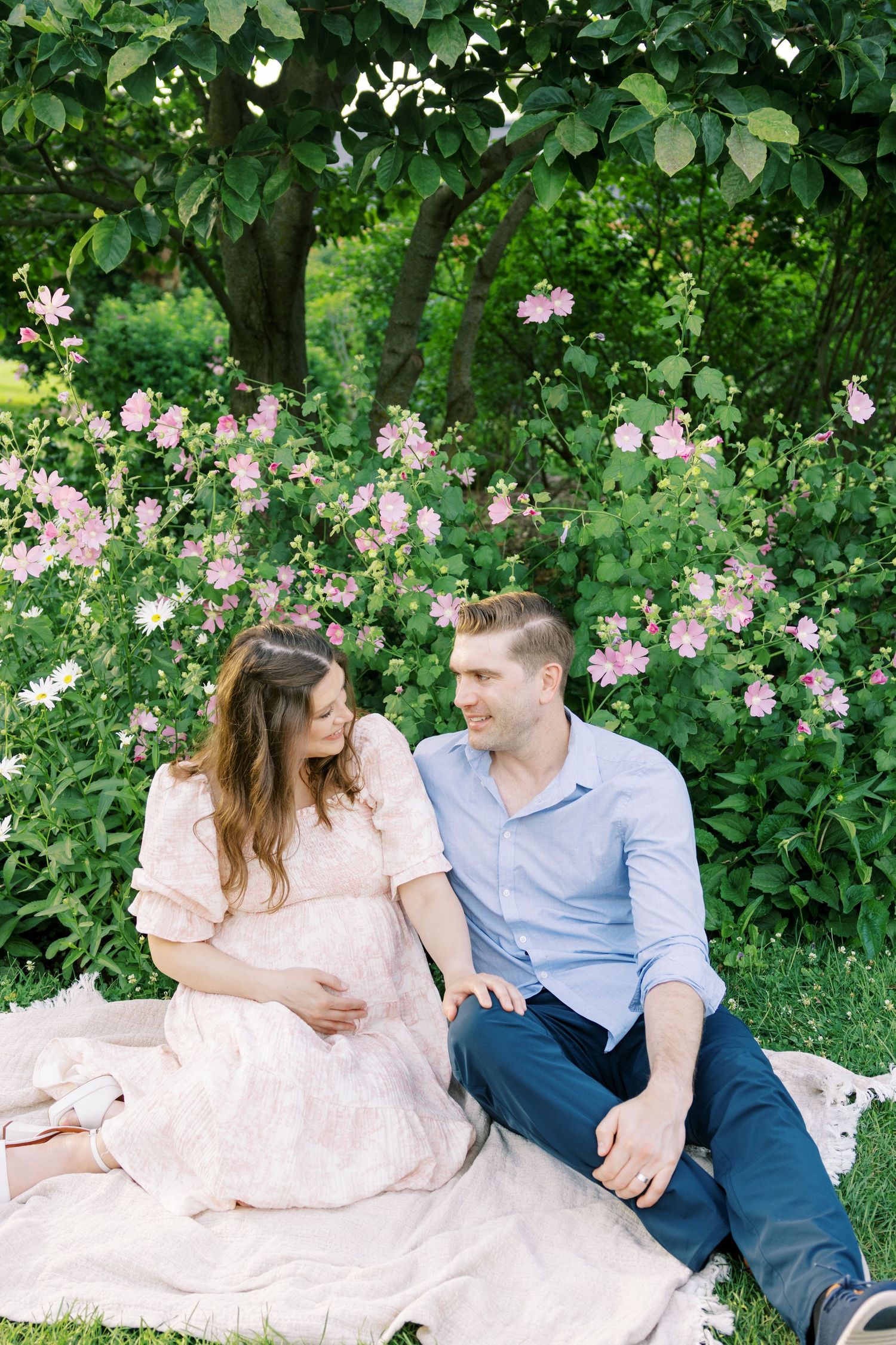 Pregnant couple sitting on blanket in lush garden with pink flowers, sharing a tender moment outdoors.