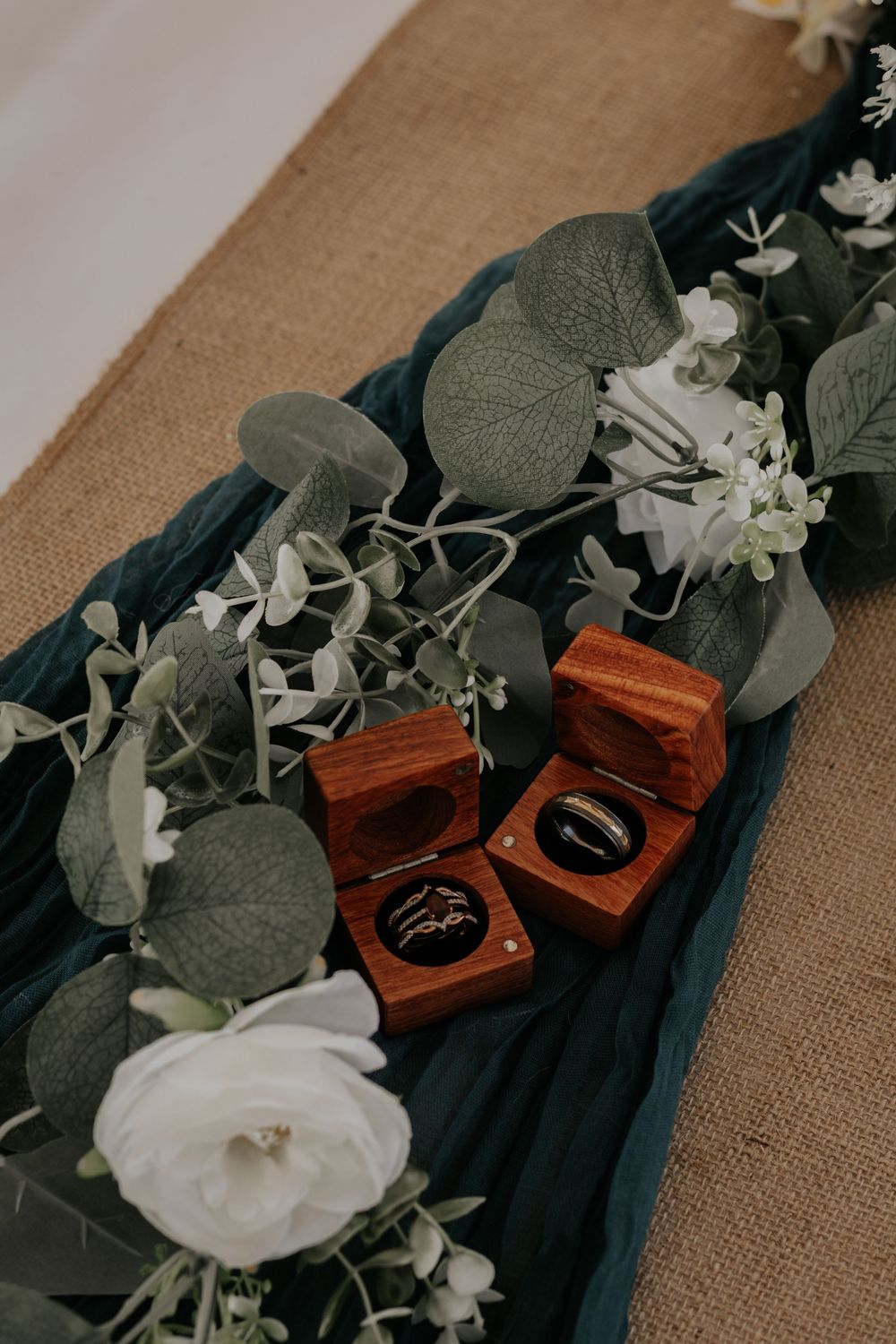 Wedding rings in wooden boxes displayed on teal fabric with eucalyptus greenery and white roses on burlap table runner.