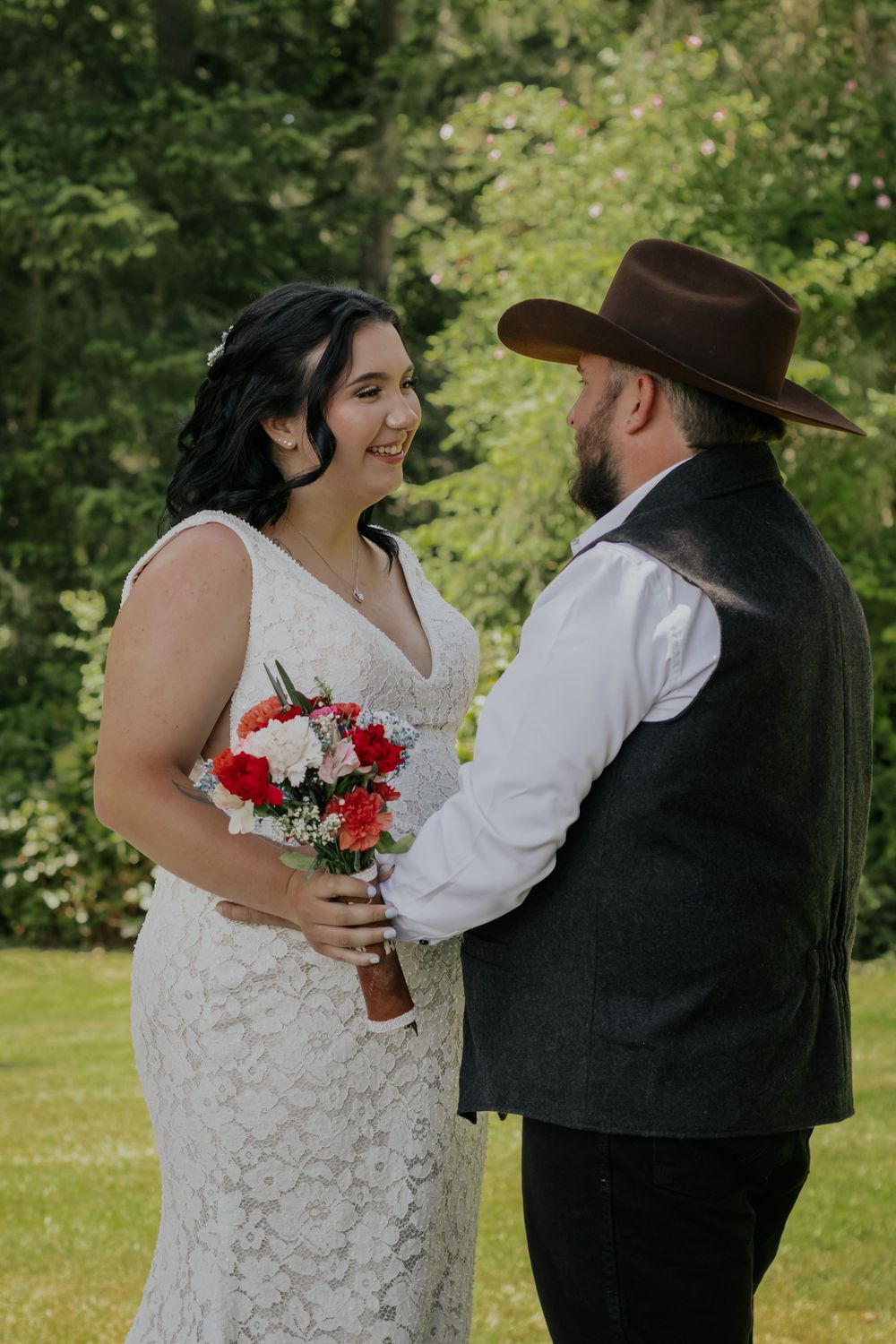 Bride in lace wedding dress holding red and white bouquet smiles at groom wearing brown cowboy hat outdoors.