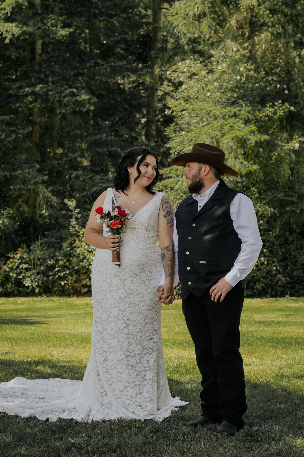 Bride in lace gown holding red bouquet stands with groom in cowboy hat and vest on green lawn with trees.