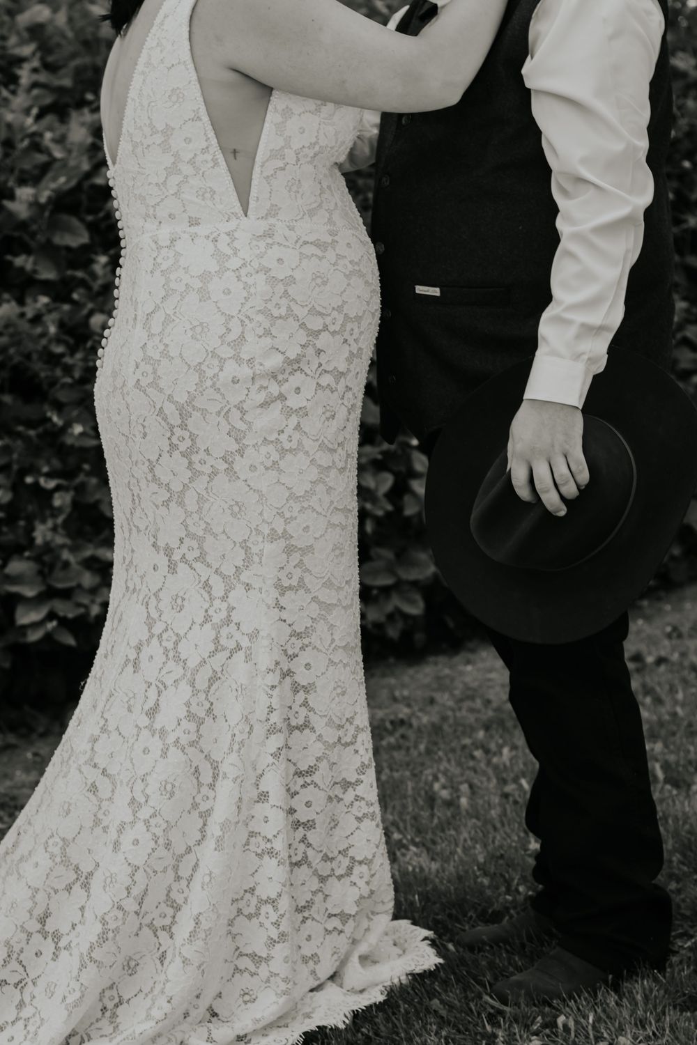 Black and white photo of a bride in lace wedding gown and groom holding cowboy hat standing together outdoors.