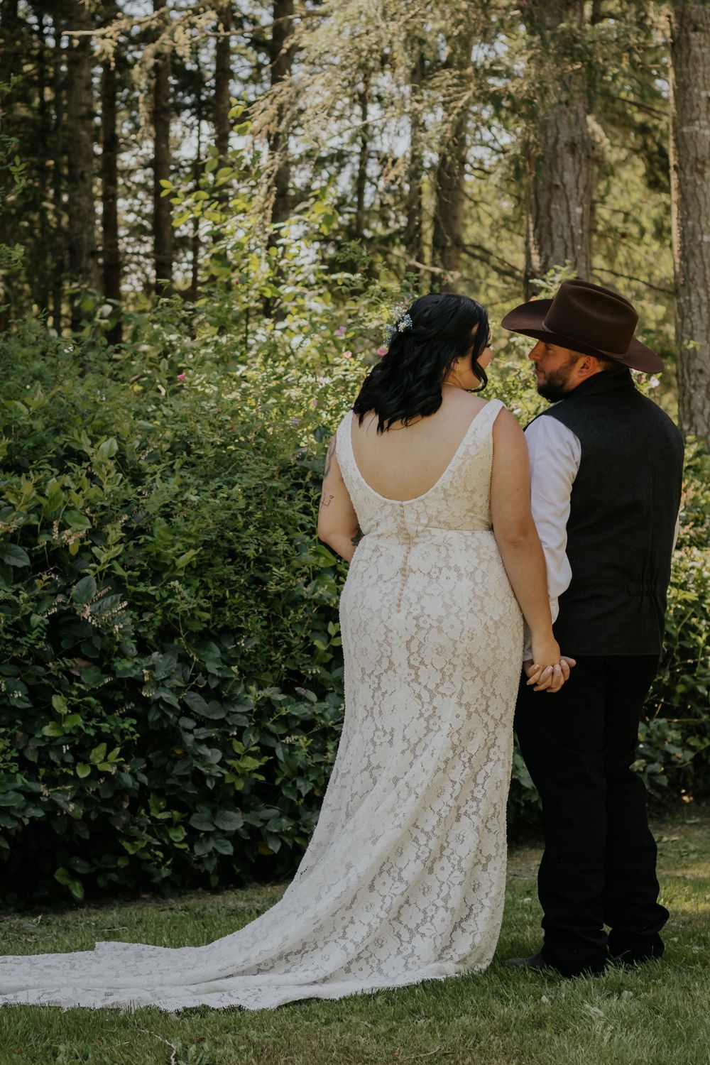 A bride in lace gown and groom in cowboy hat hold hands facing lush greenery on their wedding day.