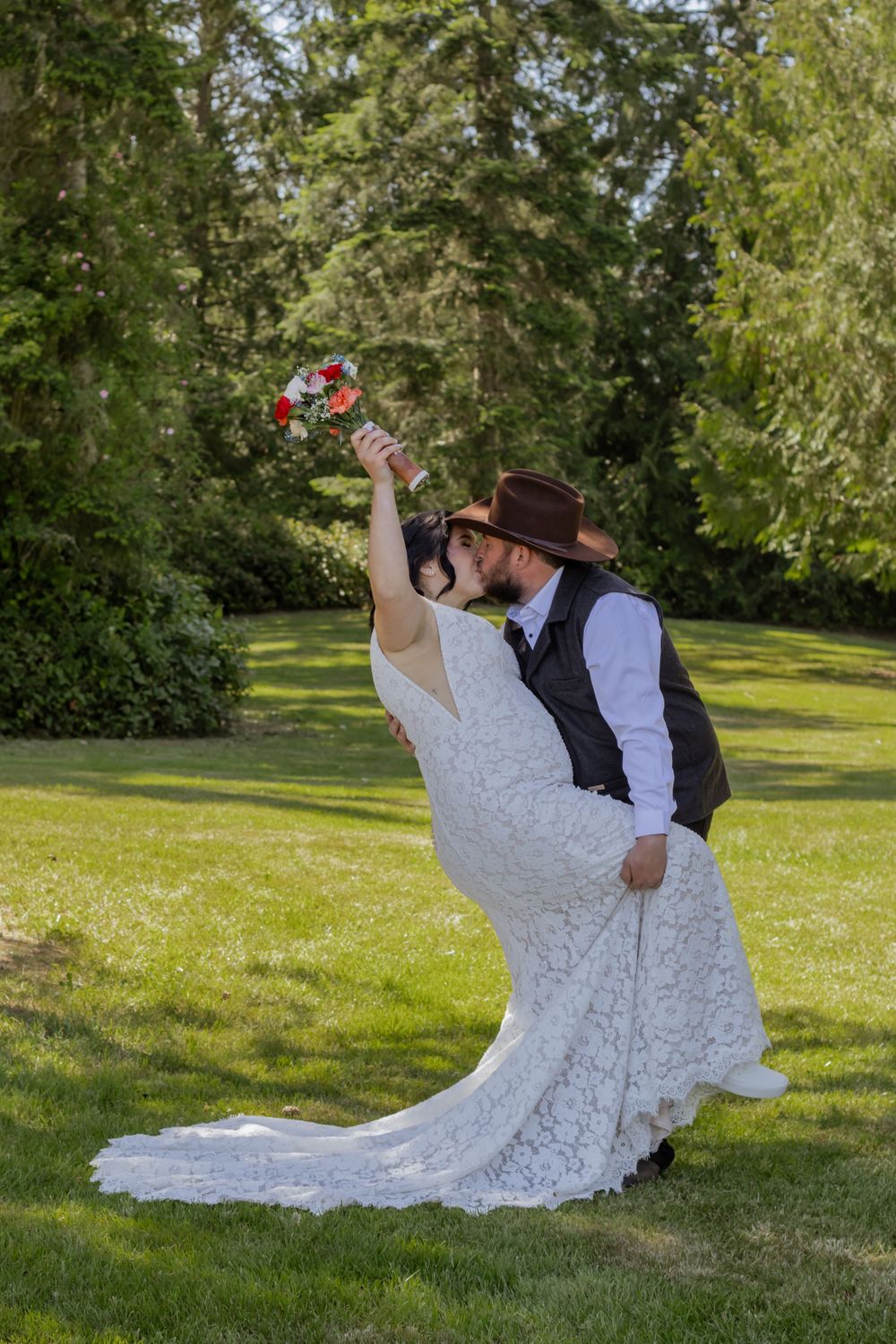 Bride and groom share a romantic dip kiss outdoors, she raises bouquet, he wears cowboy hat, lush green trees behind.