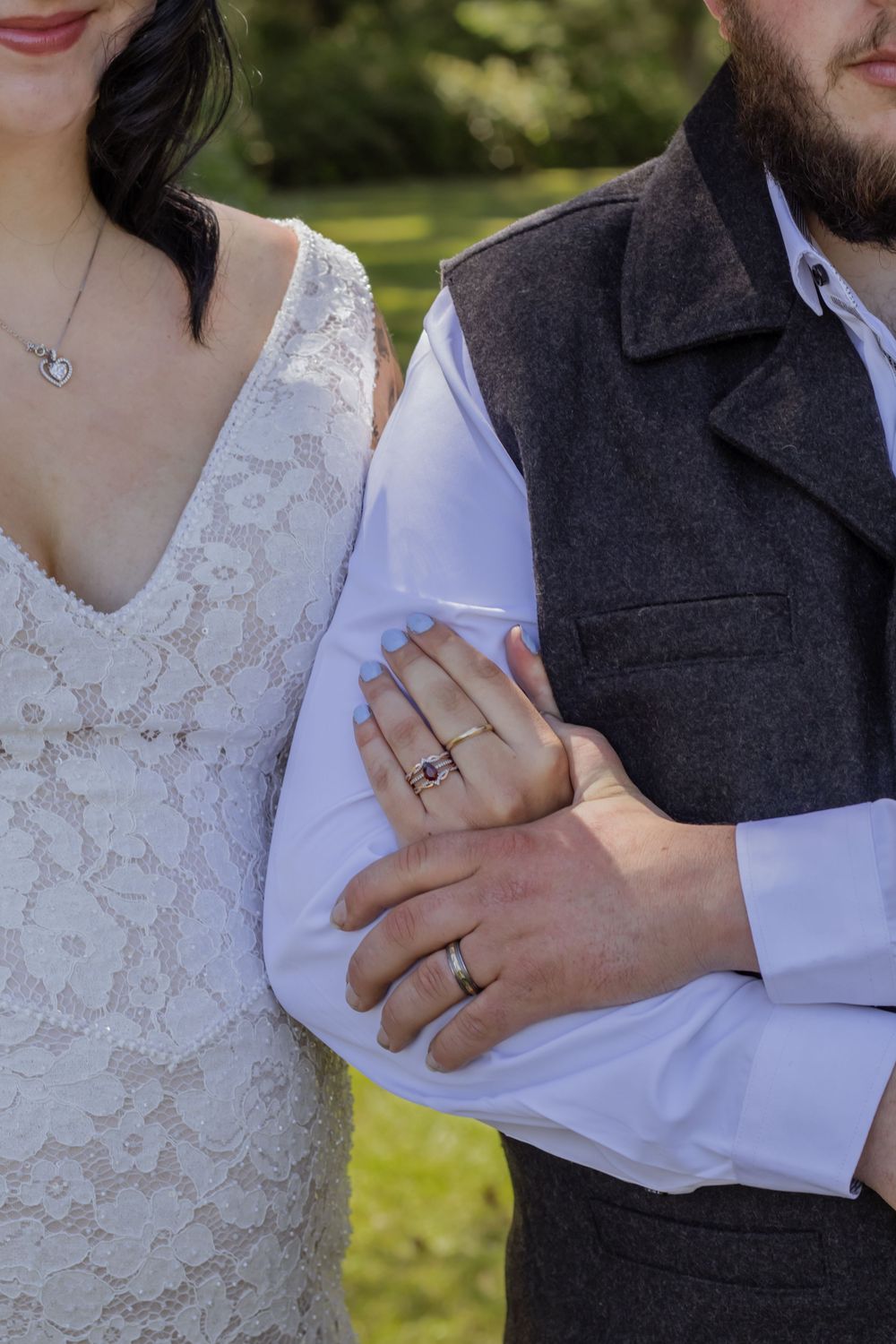 Bride and groom standing together, showcasing wedding rings, lace bridal gown, and gray vest outdoors.