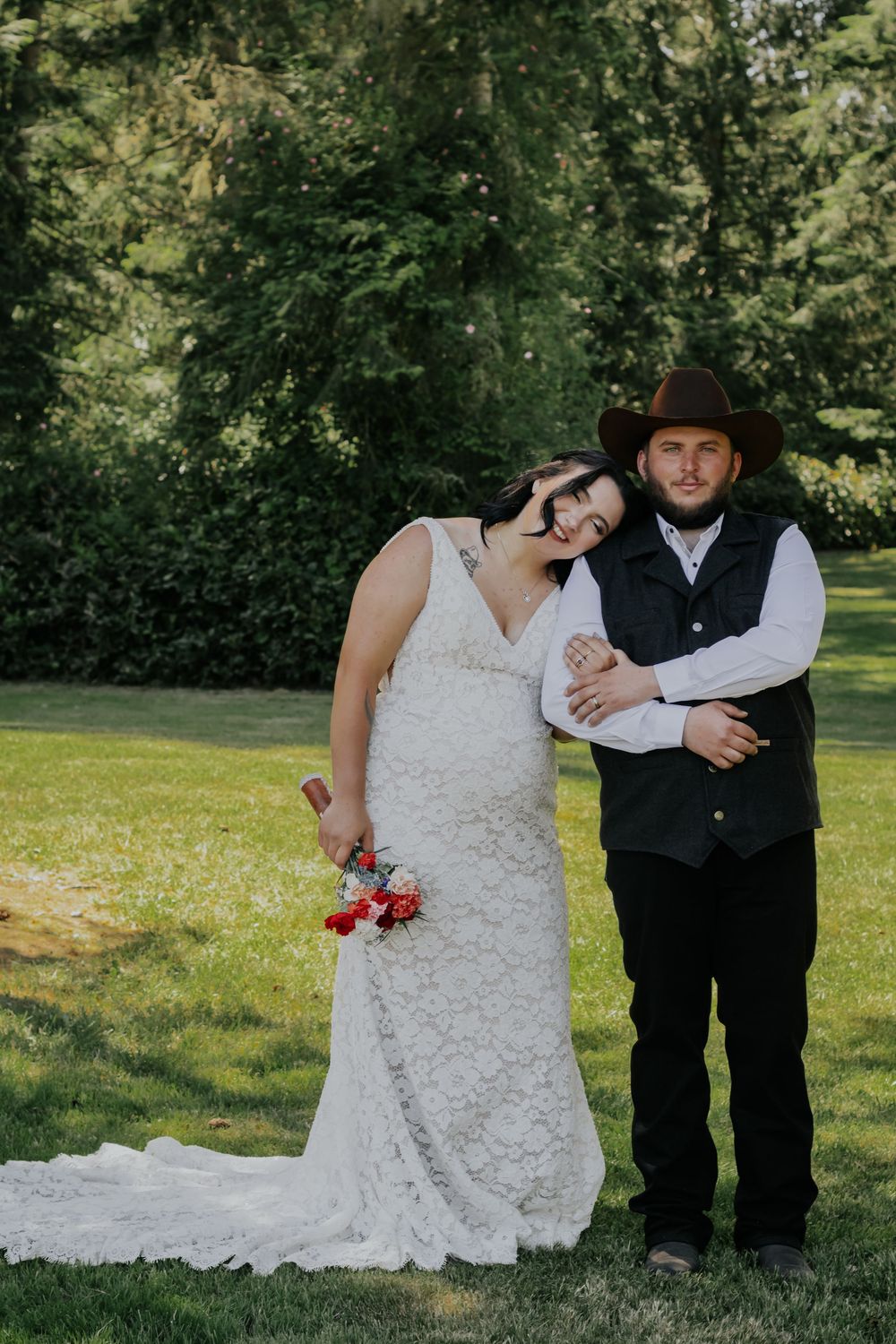 Bride in lace gown with red bouquet leans on groom in cowboy hat and black vest outdoors on green lawn.