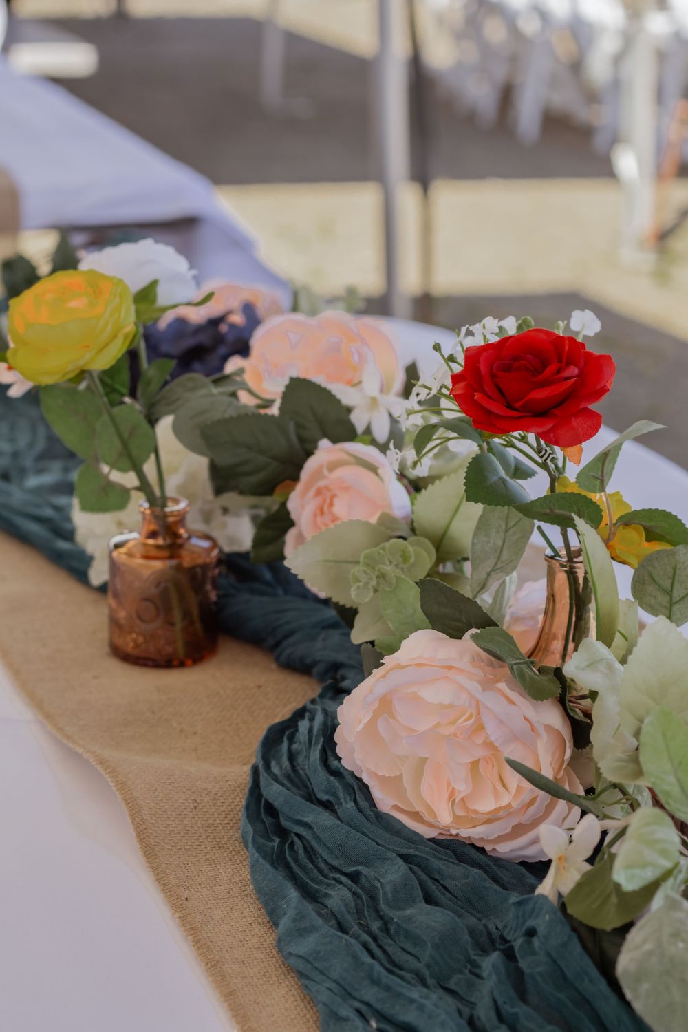 Colorful floral table runner with pink, red, and yellow roses on burlap with teal fabric at an outdoor wedding.