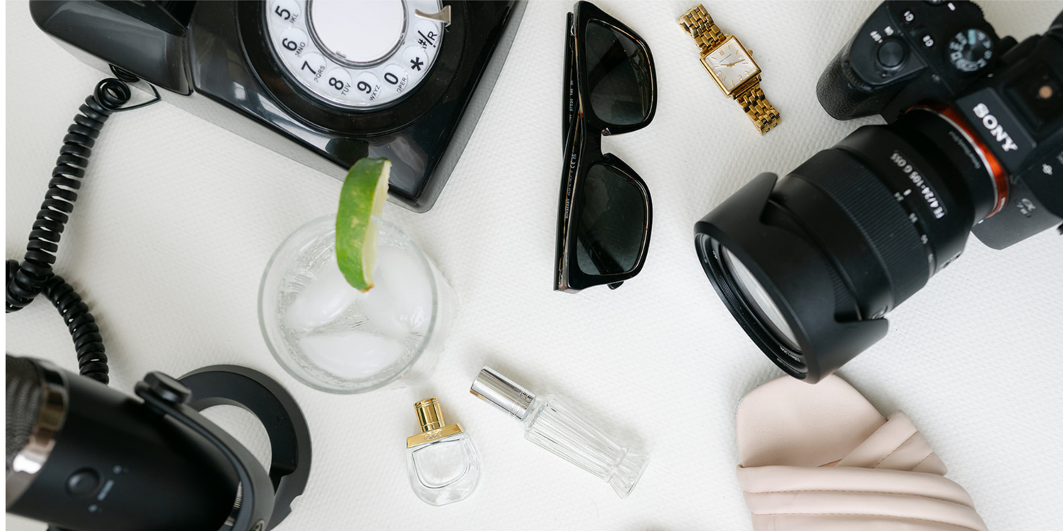 Flat lay of lifestyle items including a Sony camera, black rotary phone, microphone, sunglasses, heels, perfume, watch, and cocktail.