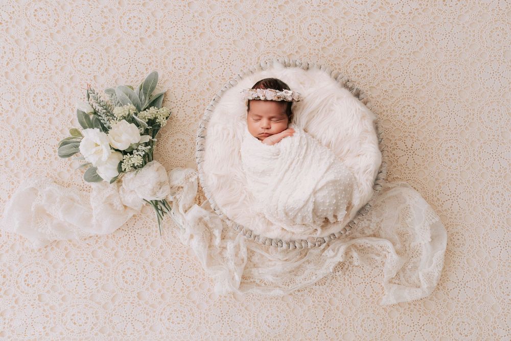 Newborn baby swaddled in white wrap sleeping in a round basket with a white floral bouquet beside her.