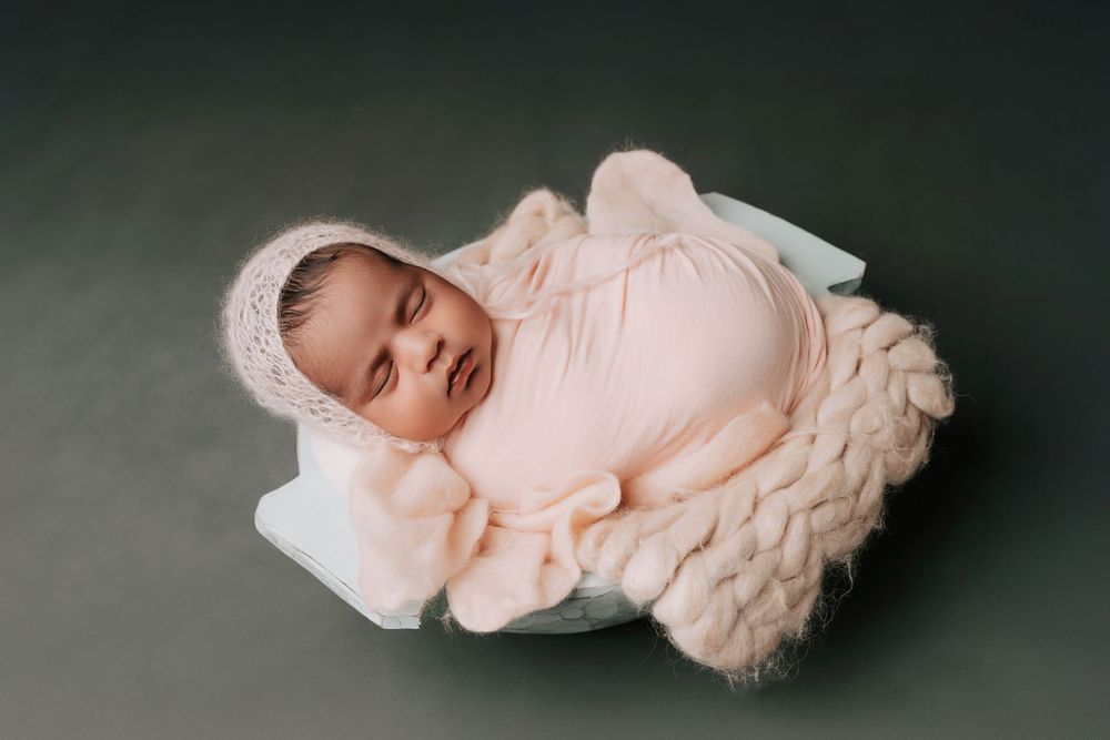 Newborn baby wrapped in pink swaddle and bonnet, sleeping peacefully in a small bowl on a green background.