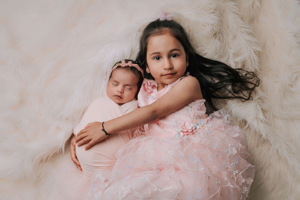 Young girl in pink dress gently holds sleeping newborn sibling on fluffy white fur backdrop.