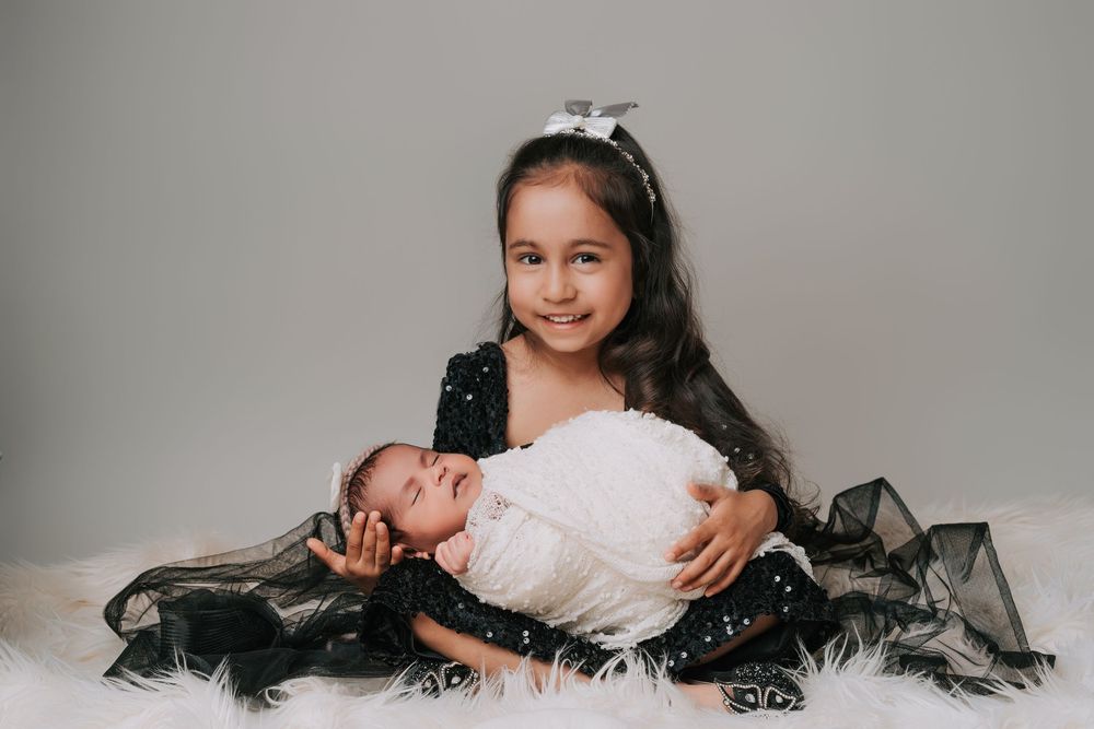 Young girl in black dress smiles holding swaddled newborn baby on white fur rug in studio portrait.