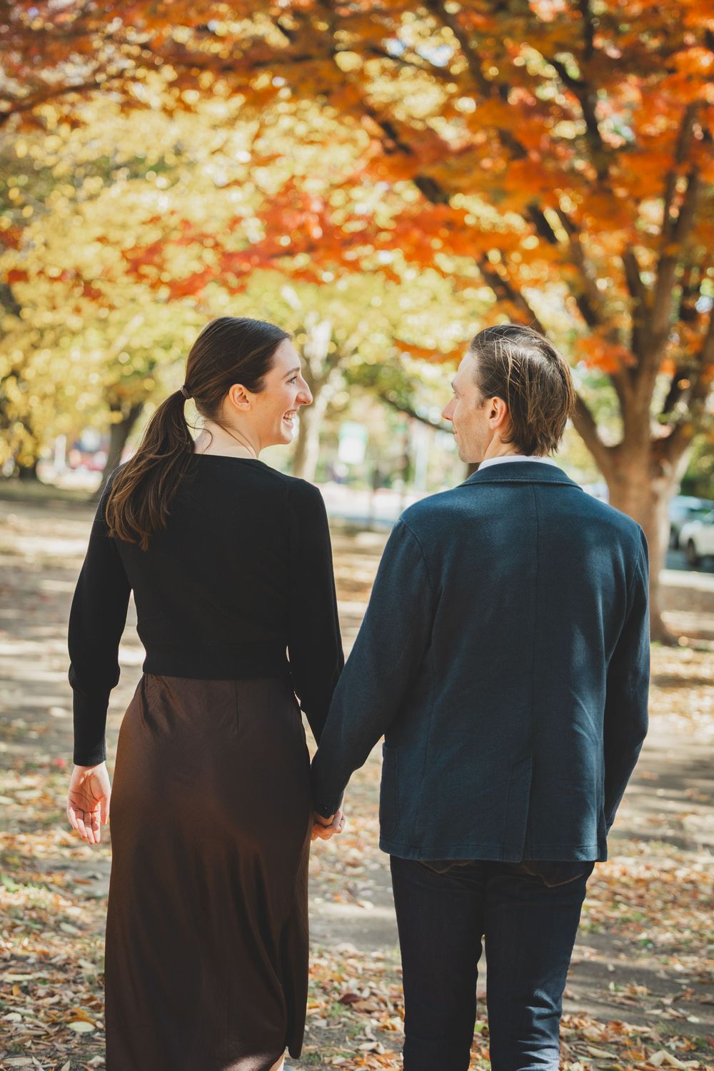 Couple holding hands walking among vibrant autumn trees with red and yellow foliage in a park.