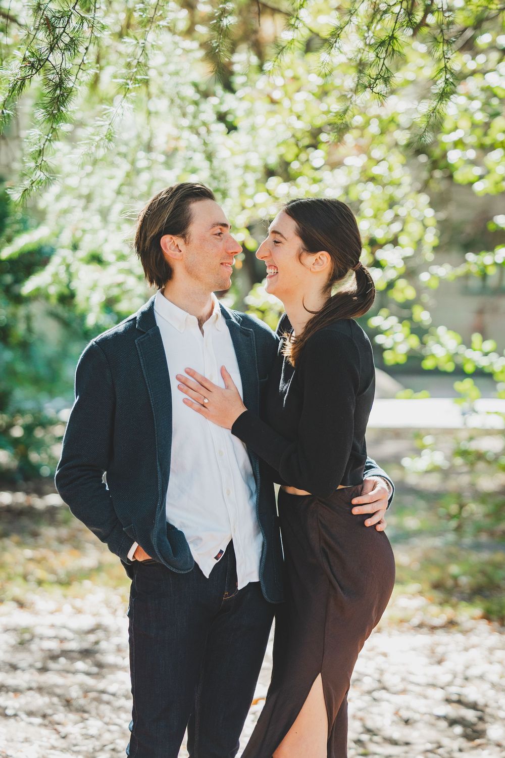 Happy couple embracing outdoors near blooming white spring trees, woman laughing in black outfit, man in navy blazer.