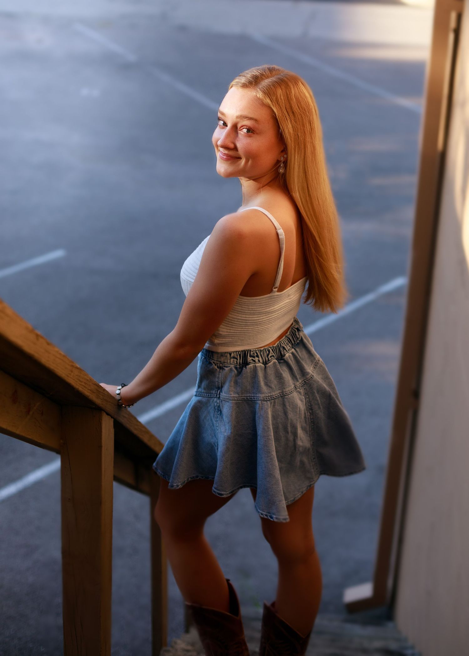 Young woman in white top, denim skirt, and brown boots poses on stairs, smiling over her shoulder.