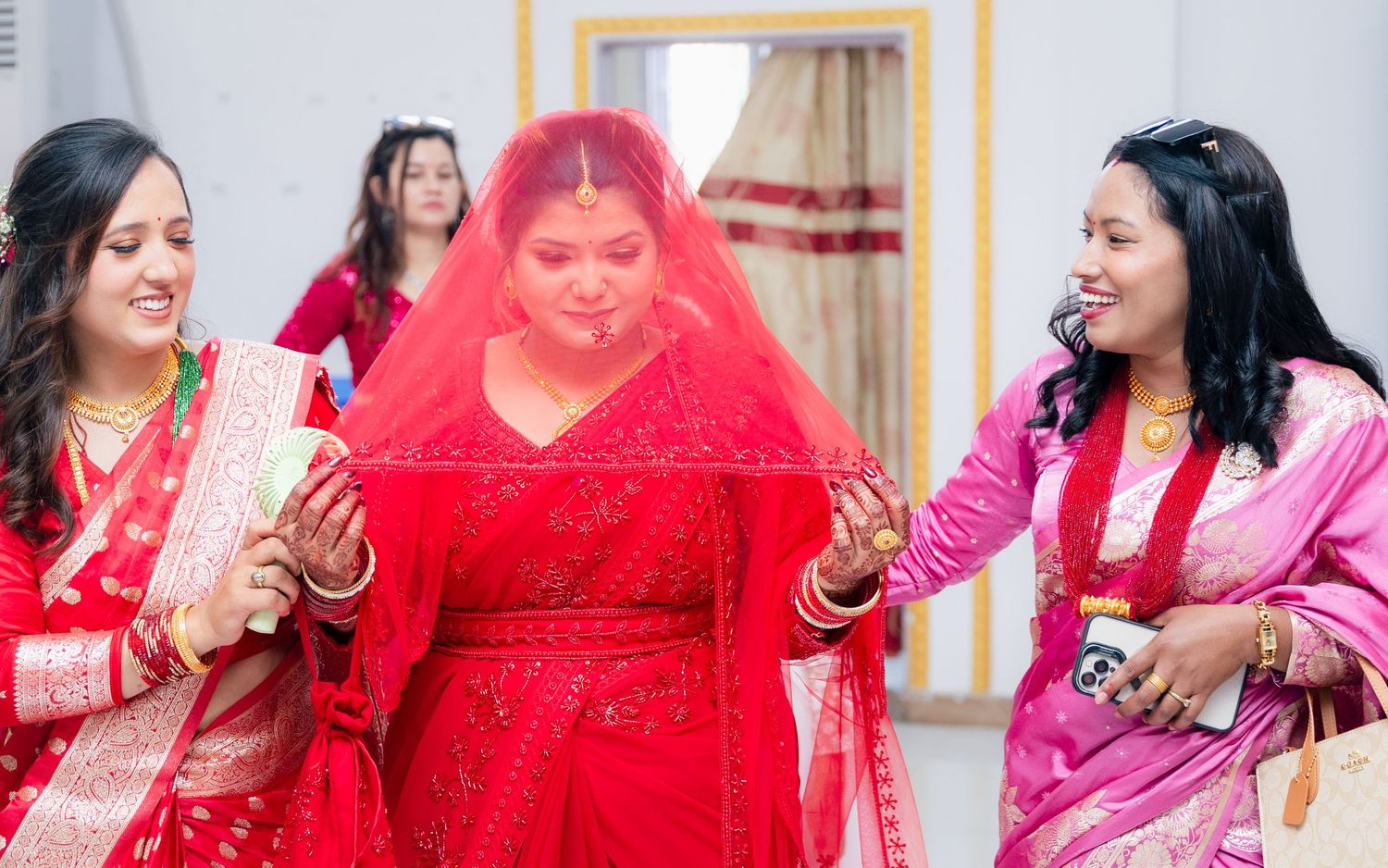 Bride in red saree and veil assisted by bridesmaids during traditional South Asian wedding ceremony.