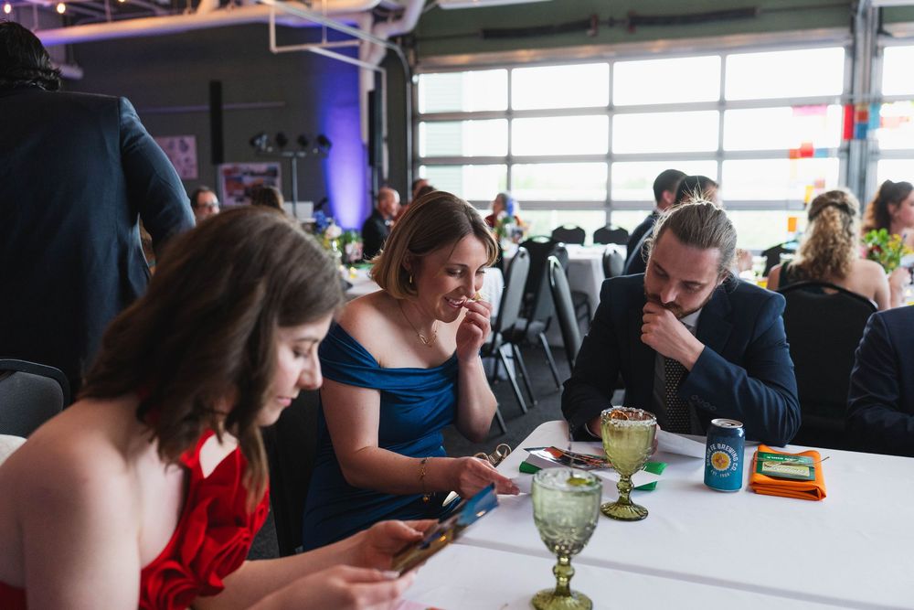Elegantly dressed guests at a formal event check phones at a white table with drinks in a bright venue.