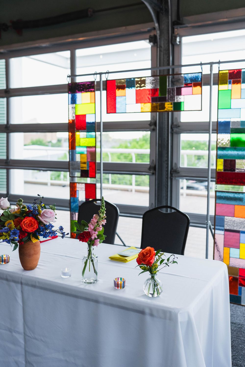 Elegant sweetheart table with white linen, floral arrangements, and colorful stained glass panels hanging in background.