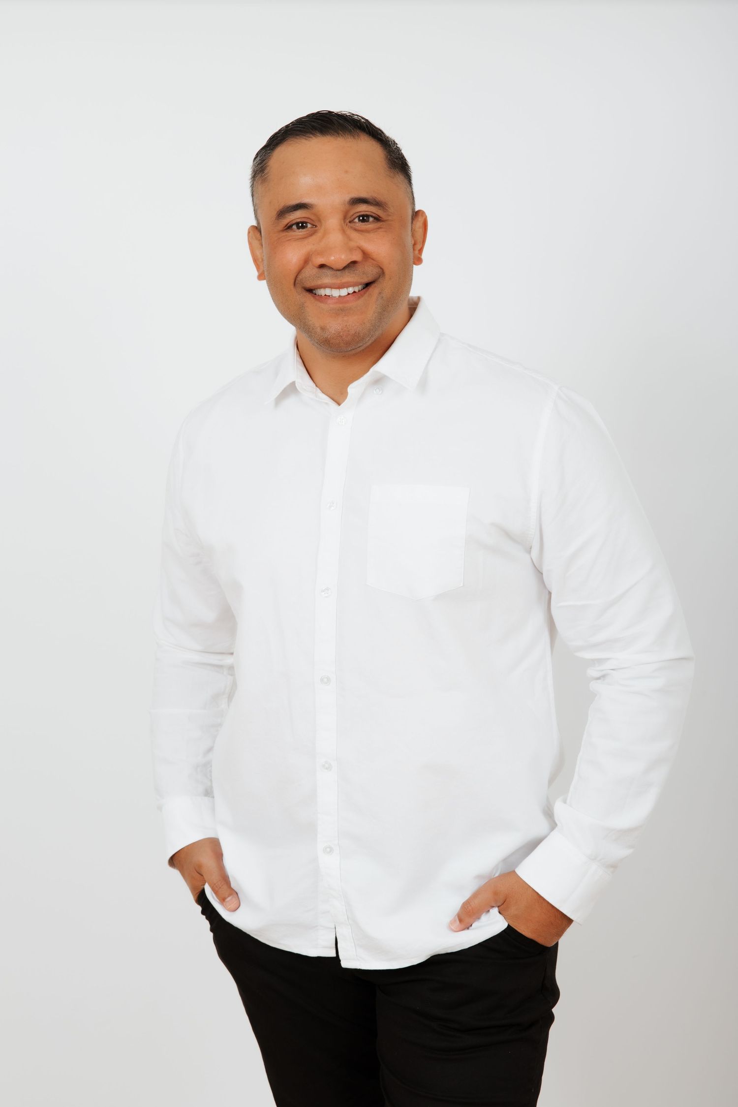 Smiling man in white button-up shirt and black pants posing with hands in pockets on white background.