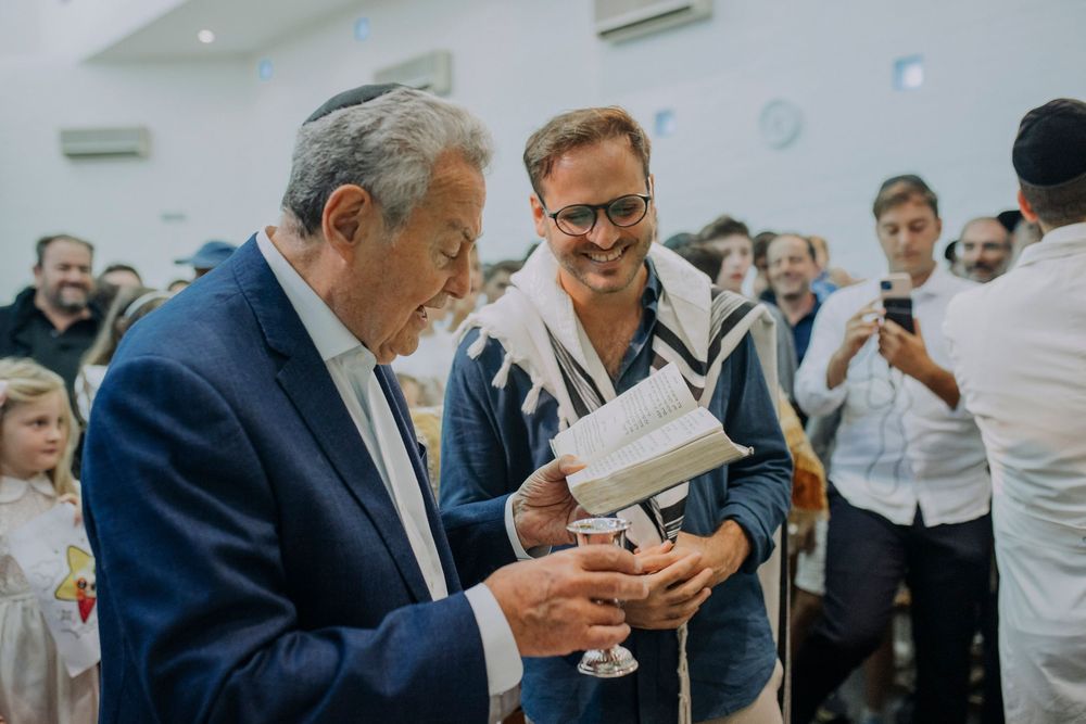 Two men in a Jewish ceremony share a meaningful moment, one wearing a tallit, holding a kiddush cup at a synagogue gathering.
