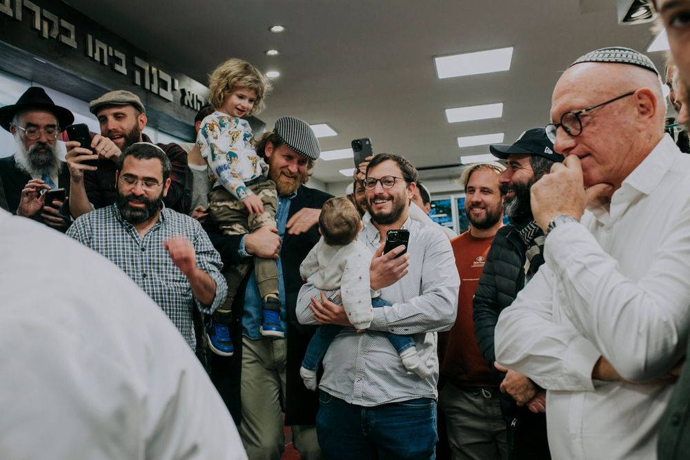 Jewish men gather joyfully at a community event, some holding children, smiling and socializing indoors.