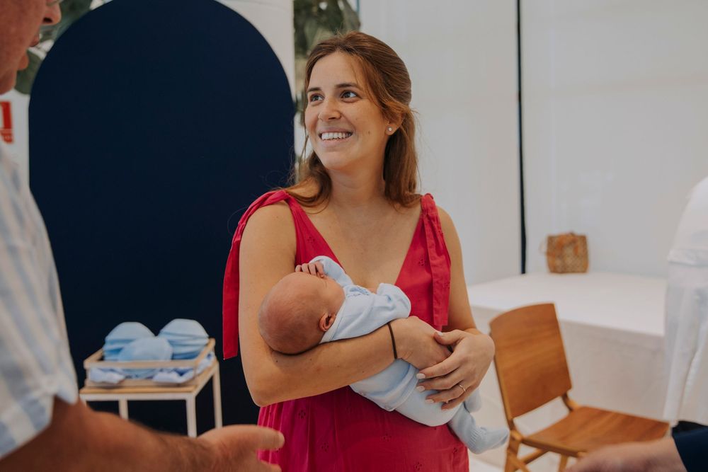 Woman in red dress holding newborn baby at a christening event, smiling warmly indoors.