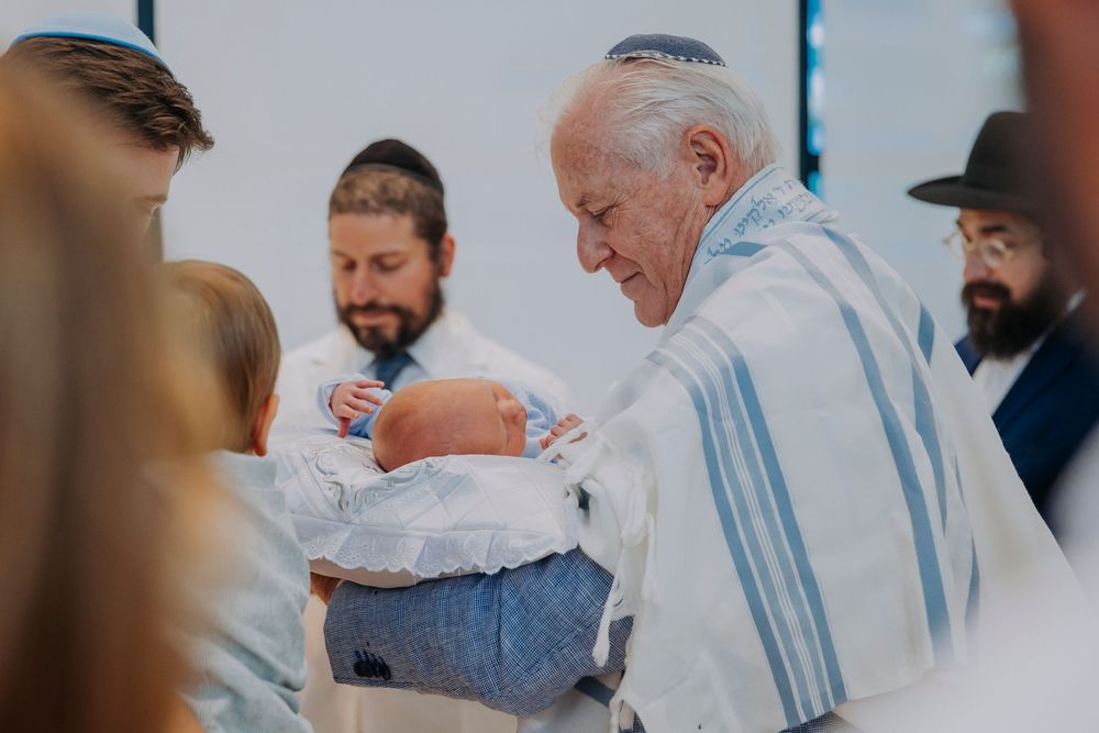 Rabbi performing a bris ceremony, holding newborn baby wrapped in white cloth during Jewish circumcision ritual.