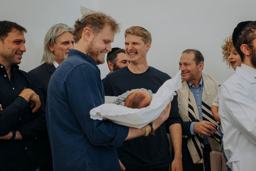 Group of men smiling at a Jewish brit milah ceremony, with one man proudly holding a newborn baby.