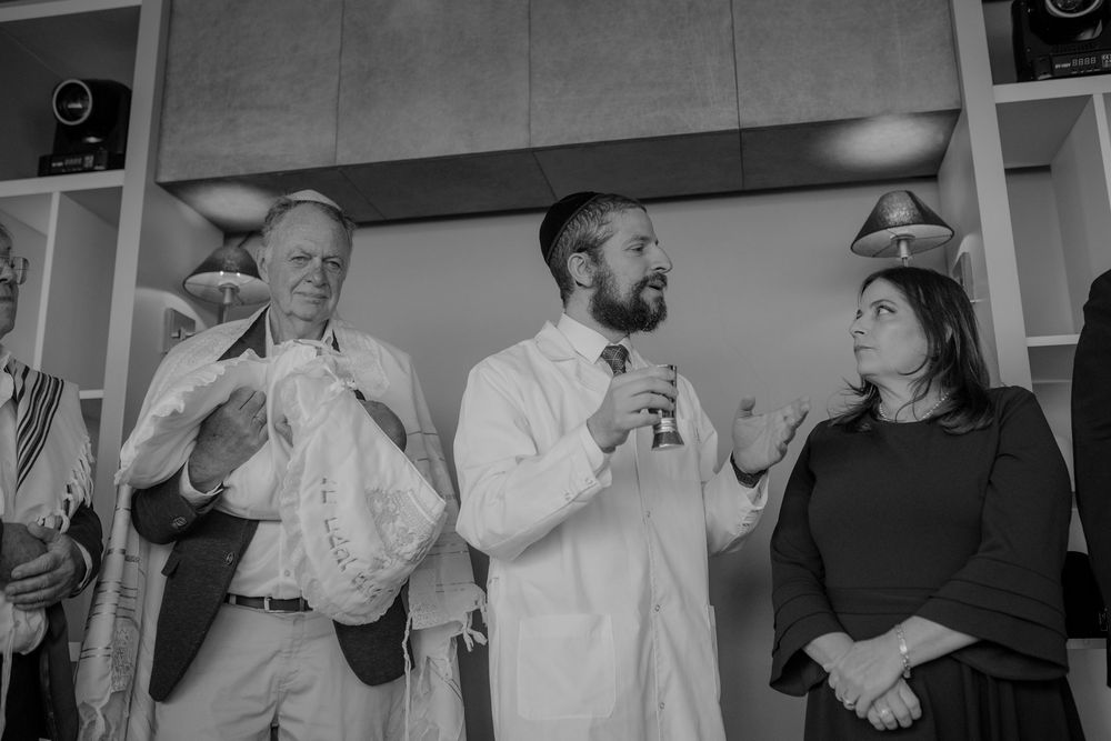 Three people stand indoors, one in a white coat speaking into a microphone during a formal ceremony.