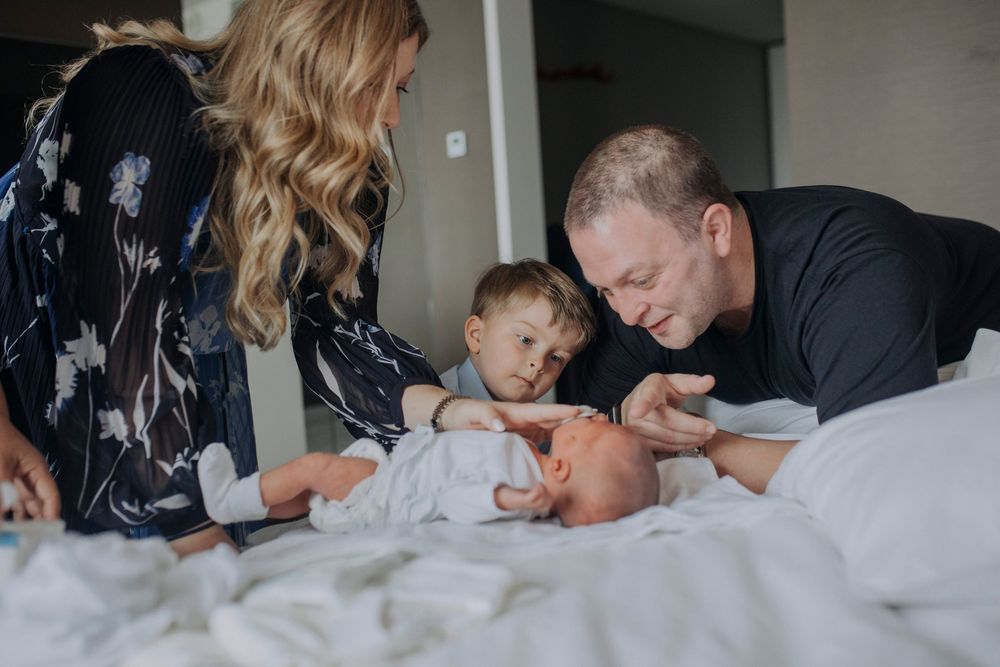 Family of four with parents and toddler lovingly gazing at newborn baby lying on white bed.