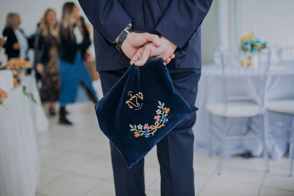 Man in blue suit holding navy embroidered velvet bag with floral and dove design at elegant wedding reception.