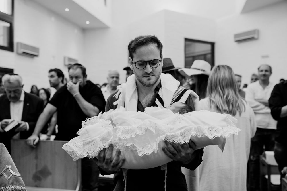 Man in tallit holding a baby wrapped in white lace during a Jewish religious ceremony in a synagogue.