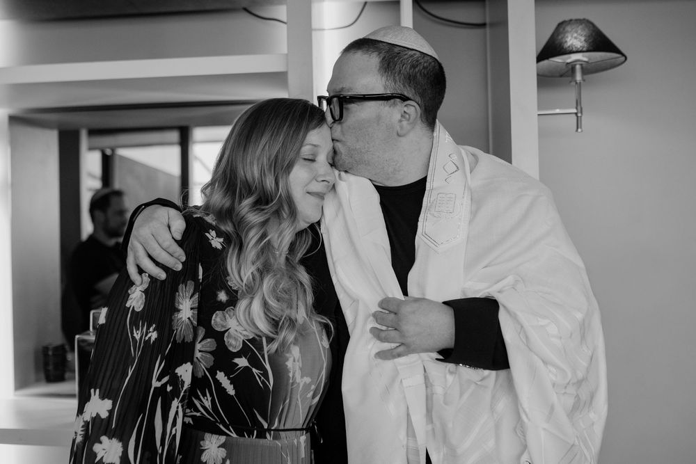 Black and white photo of a man in white robe kissing a smiling woman on the cheek indoors.
