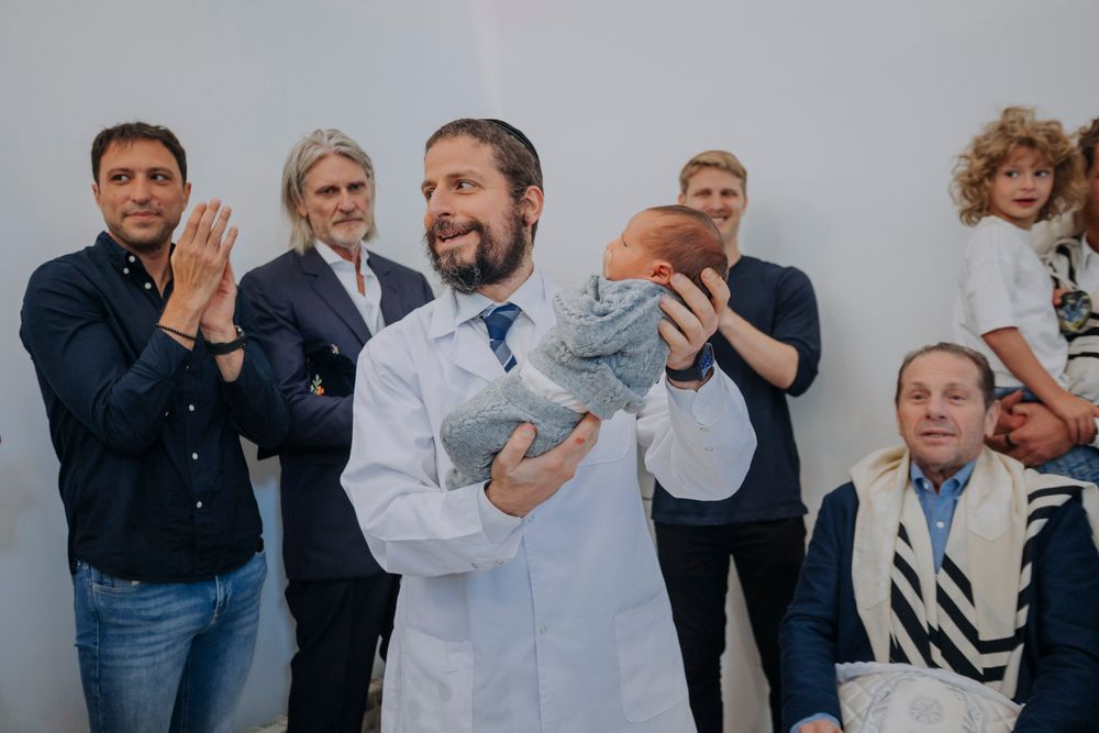 Rabbi in white robe holds newborn baby at a Jewish bris ceremony surrounded by smiling family and guests.