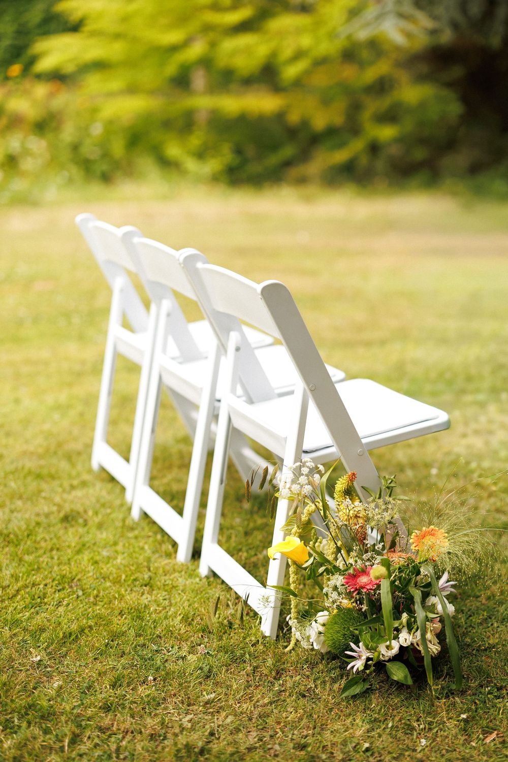 White folding chairs with colorful floral arrangement on green grass at an outdoor wedding ceremony.