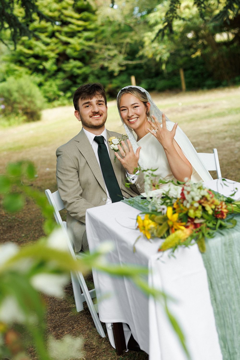 Bride and groom sit at outdoor wedding table, smiling as bride shows off her ring, surrounded by lush greenery.