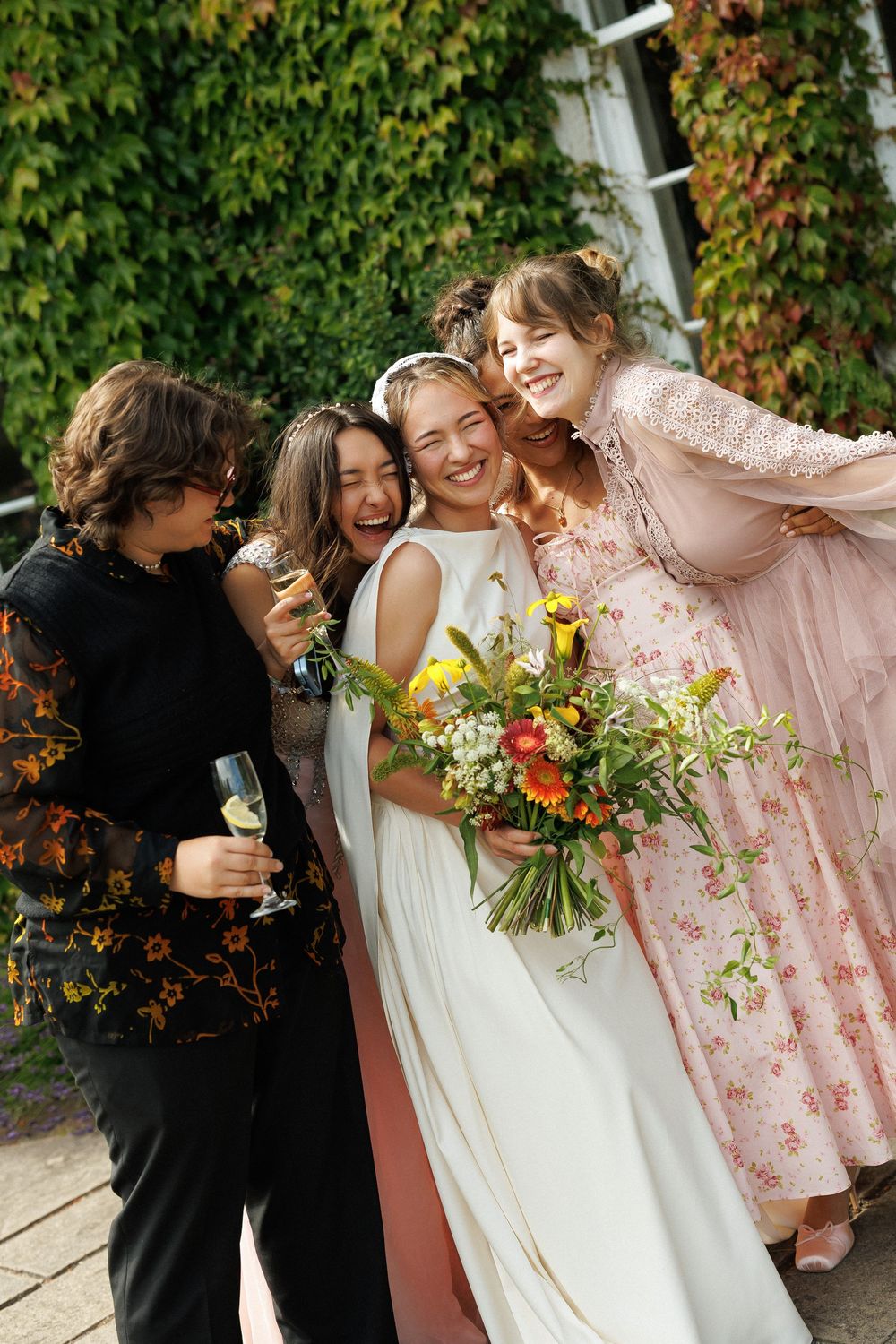 Bride in white gown holds colorful bouquet, laughing with bridesmaids in pink and black outside ivy-covered building.