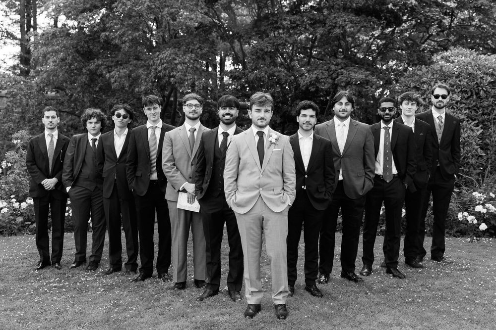 Black and white photo of a groom and groomsmen in suits posing outdoors in a garden setting.