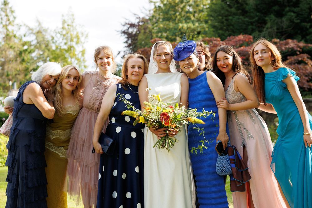 Bride with colorful bouquet poses with wedding guests in elegant dresses outdoors in a sunny garden.