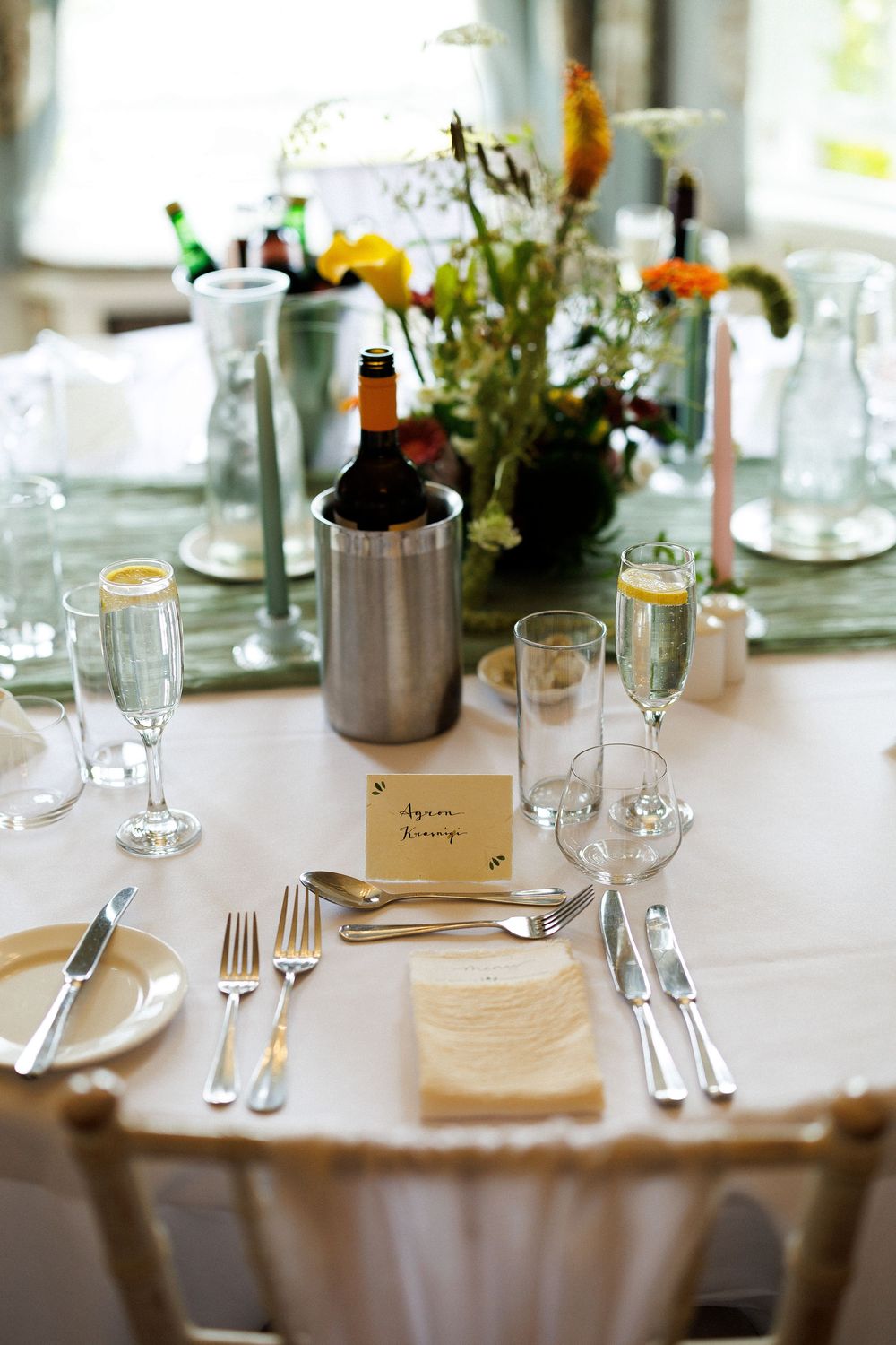 Elegant wedding reception table setting with champagne flutes, silverware, wine bucket, floral centerpiece, and place card.