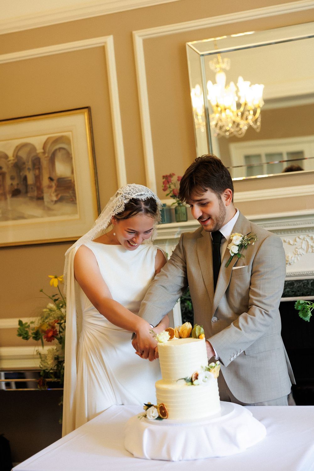 Bride and groom smiling while cutting a two-tier white wedding cake decorated with flowers at their elegant reception.