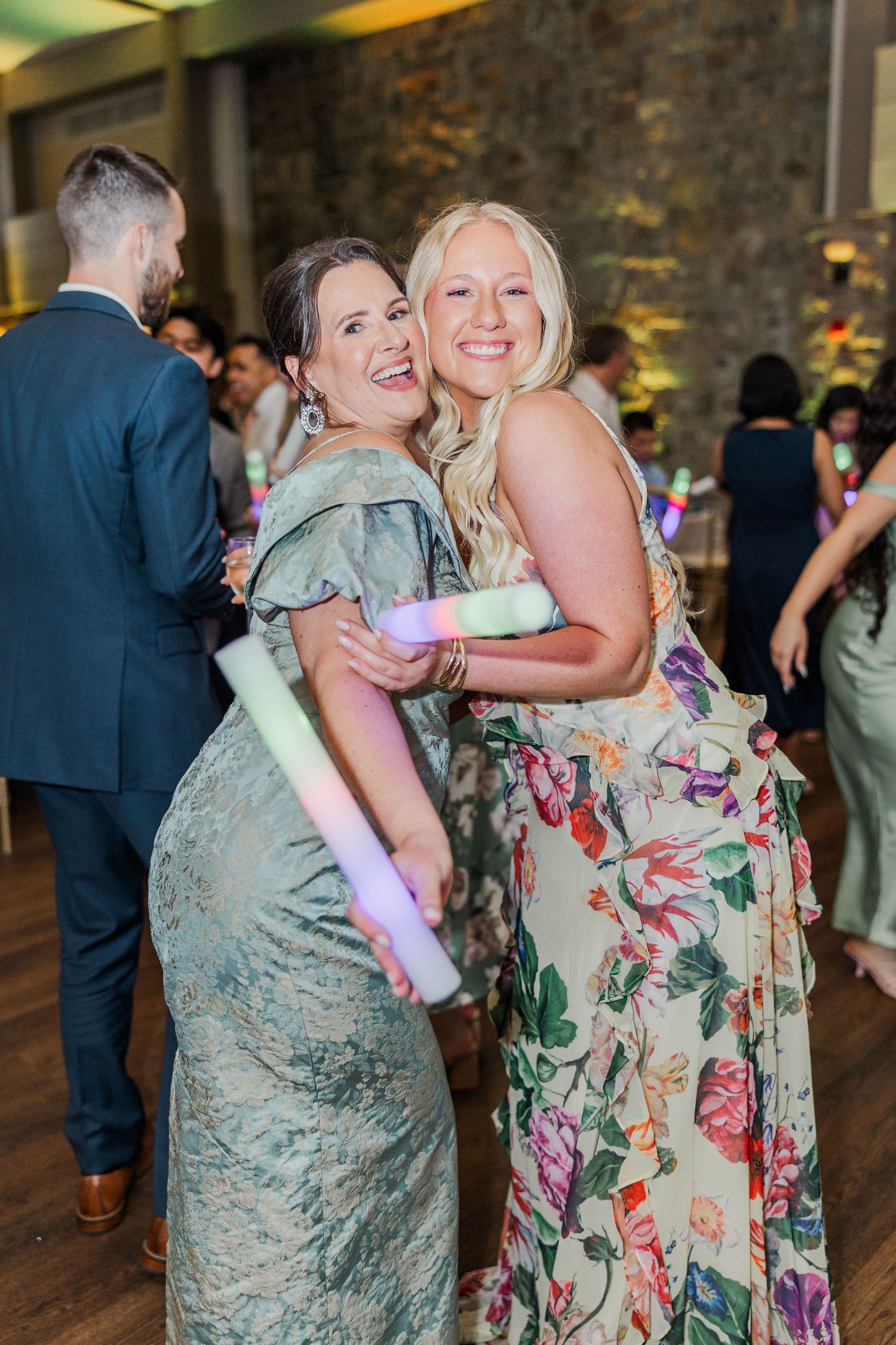 Two women in elegant dresses smile and hug while holding glow sticks at a wedding reception.
