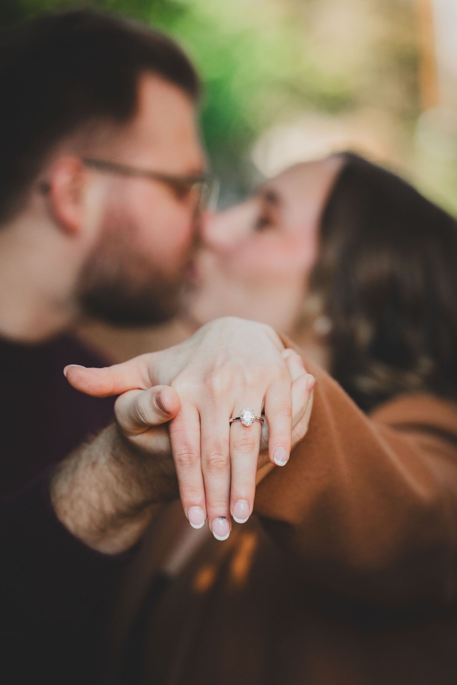 Oval diamond engagement ring in sharp focus with the couple kissing softly together in the blurred background