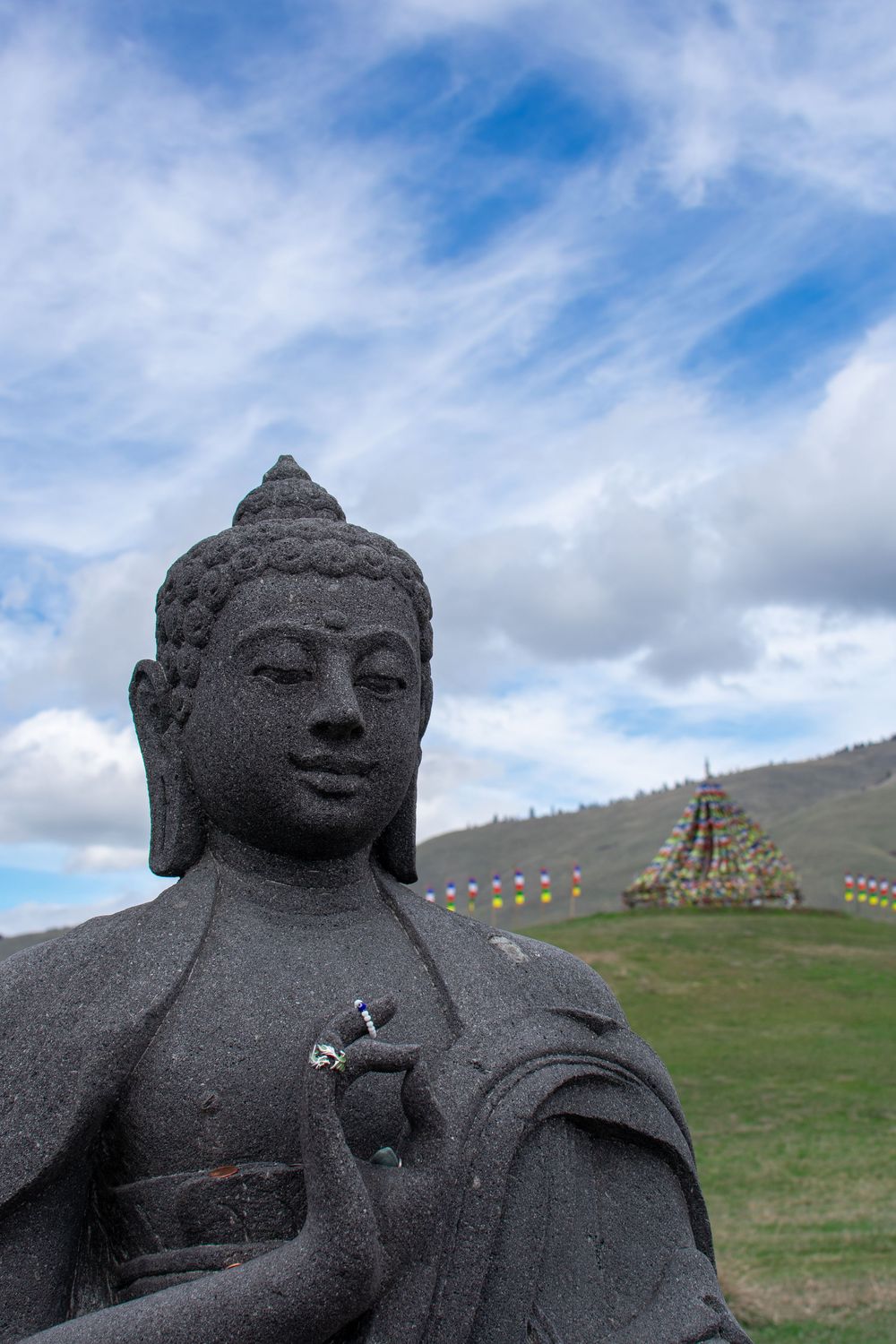 Stone Buddha statue with prayer flags and colorful stupa on green hillside under blue sky.
