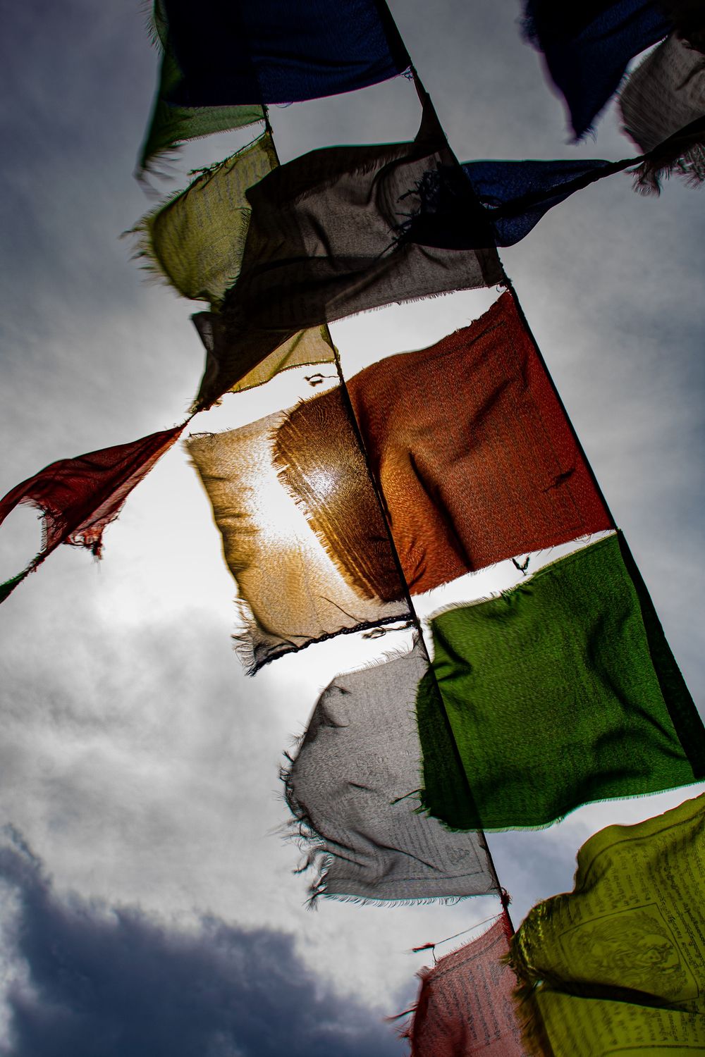 Colorful Buddhist prayer flags waving in wind against dramatic cloudy sky backlit by sun.