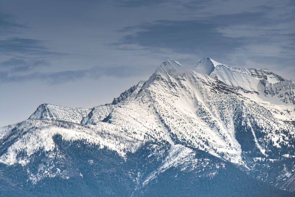Snow-capped mountain peaks rise dramatically against a blue cloudy sky in a winter alpine landscape.