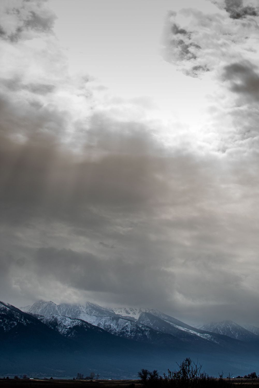 Dramatic storm clouds gather over snow-capped mountain peaks with dark skies and a faint sun breaking through.